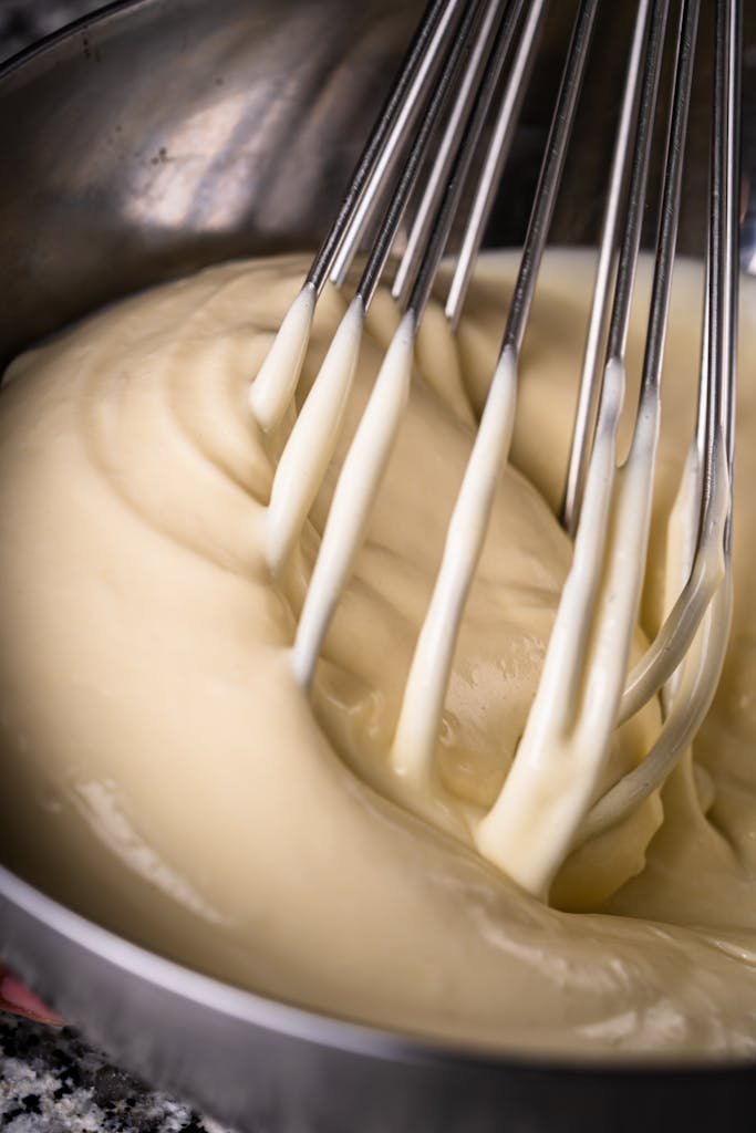 Metal whisk blending creamy batter in stainless steel bowl, ideal for baking content.